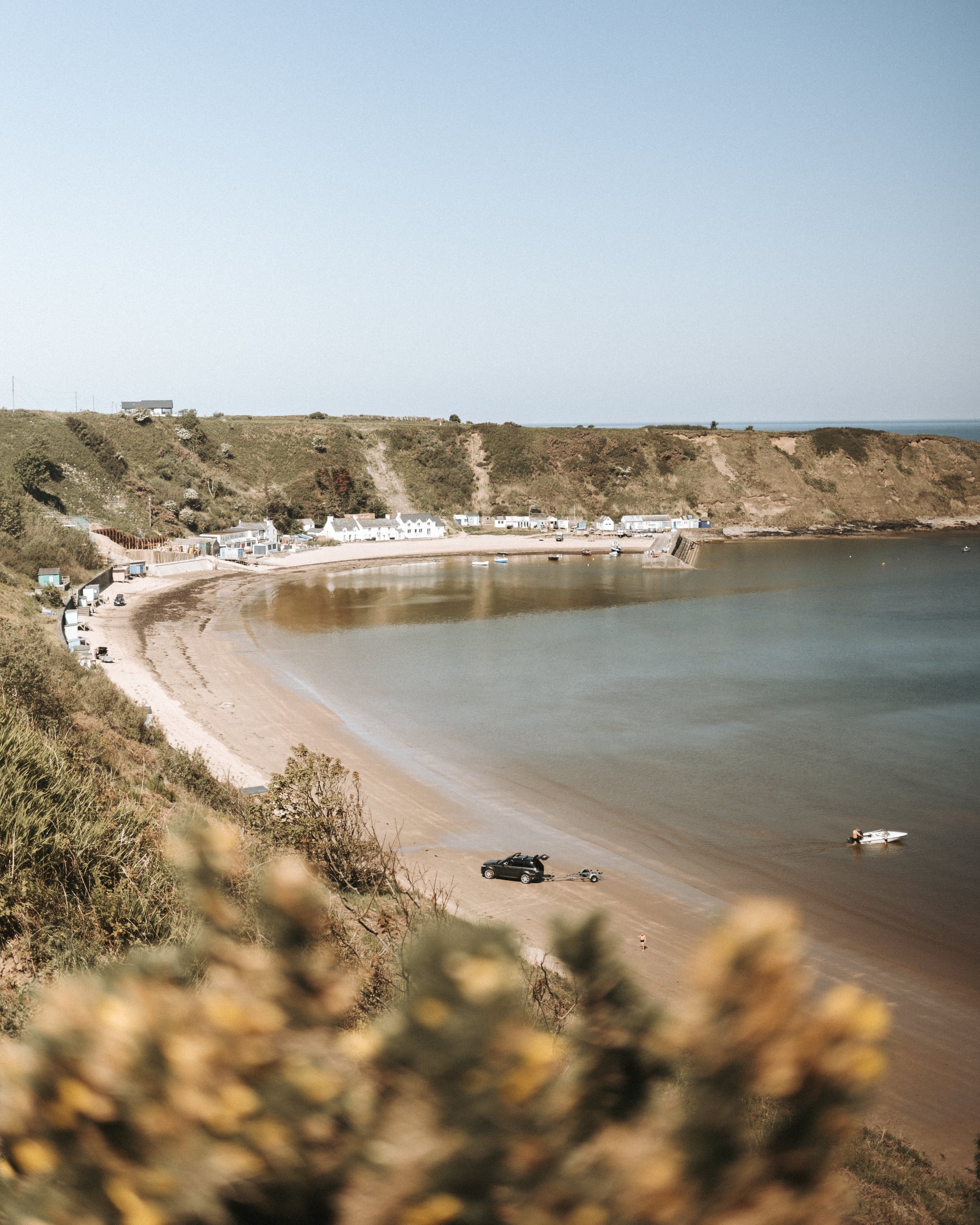 Sweeping sandy bay on the Llŷn Peninsula with whitewashed cottages, green cliffs, and a 4x4 launching a boat into calm shelt…