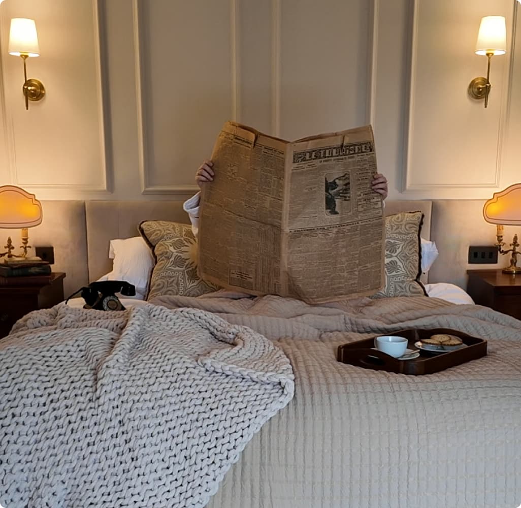 Guest reading a broadsheet newspaper in a luxury double bedroom with chunky knit throw, breakfast tray, and warm brass wall…
