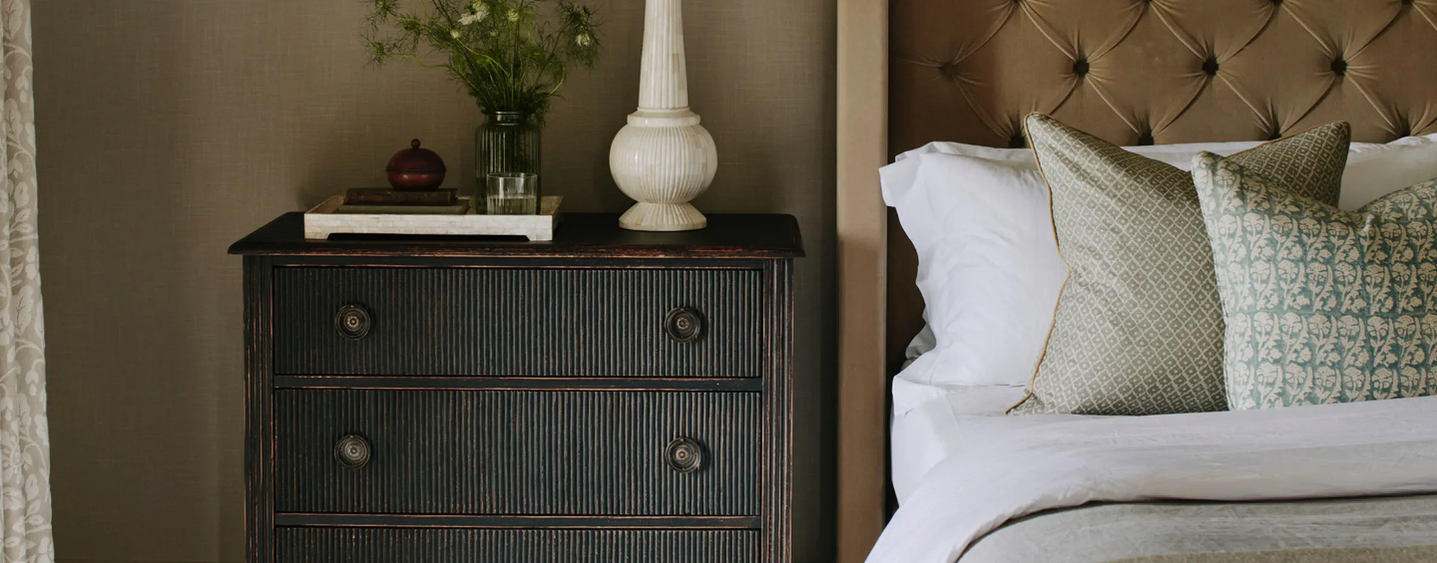 Luxury holiday let bedroom detail with tufted velvet headboard, crisp white linen, patterned cushions, and an ebonised bedsi…