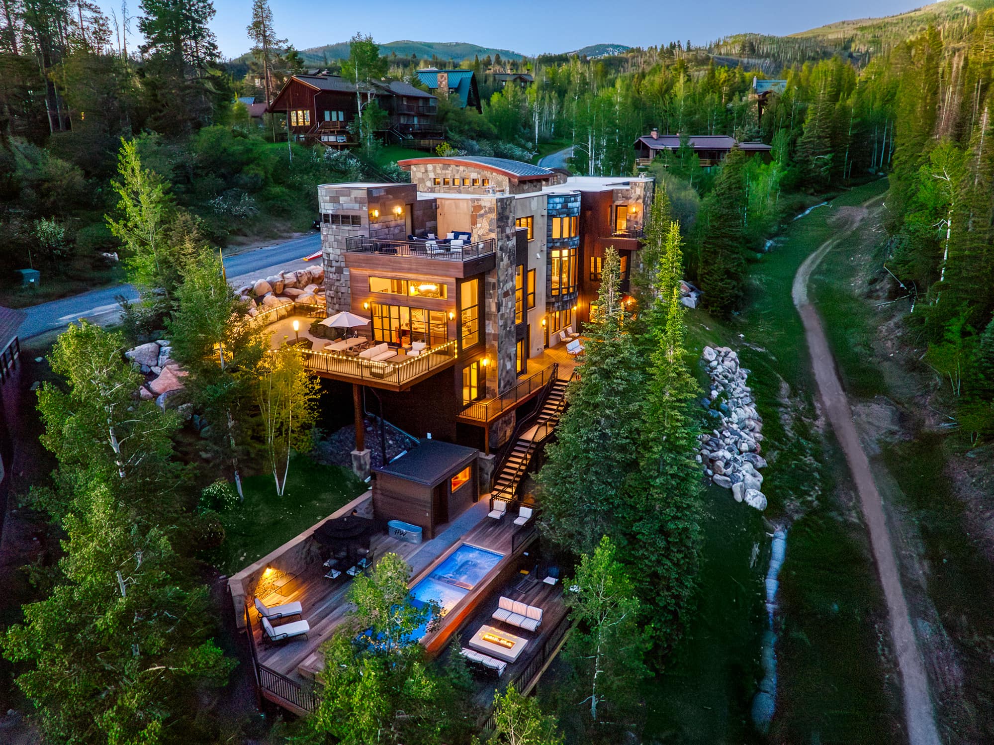 Aerial dusk view of a multi-storey luxury mountain lodge with stone facade, glowing floor-to-ceiling windows, rooftop terrac…
