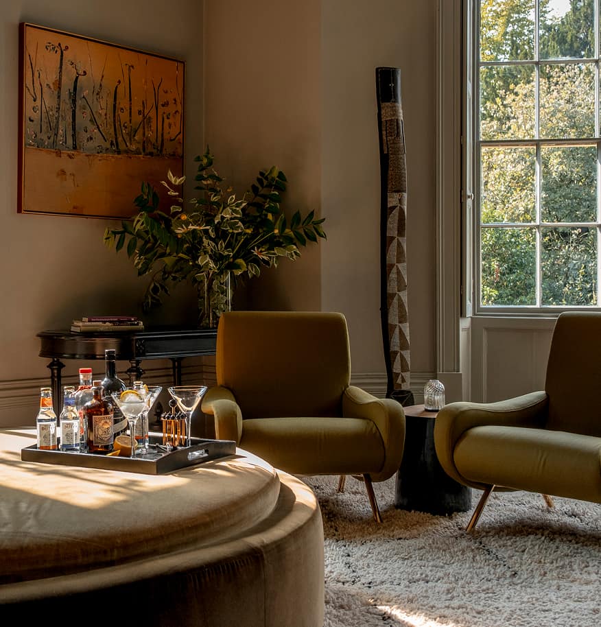 Sunlit sitting room corner with olive velvet armchairs, a drinks tray of spirits and cocktail glasses on a round ottoman, an…