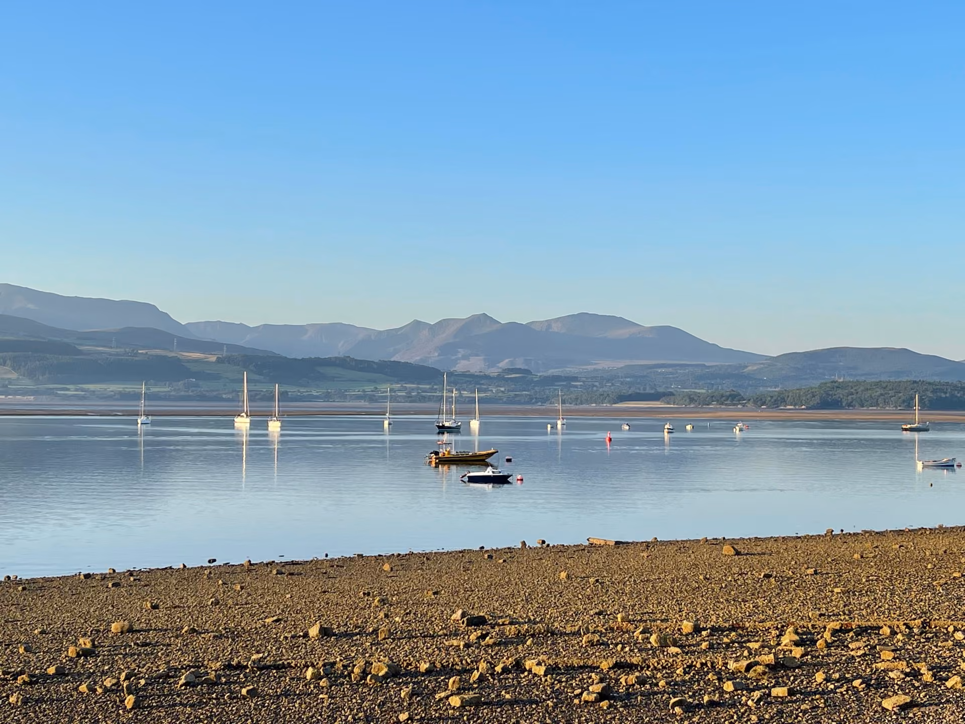 Calm estuary at low tide with sailing boats moored on glassy water and Snowdonia mountains in the distance, Anglesey coastli…