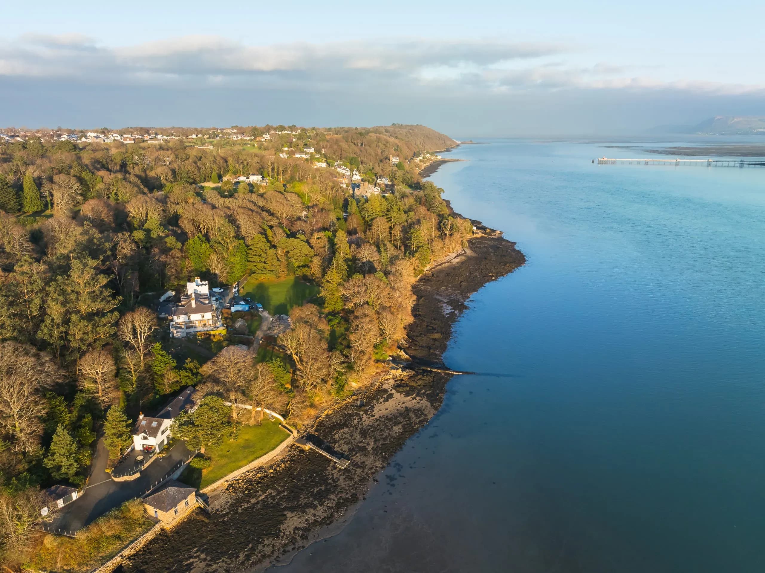 Aerial view of a white waterfront holiday let nestled among woodland on the Menai Strait coastline, with private grounds lea…