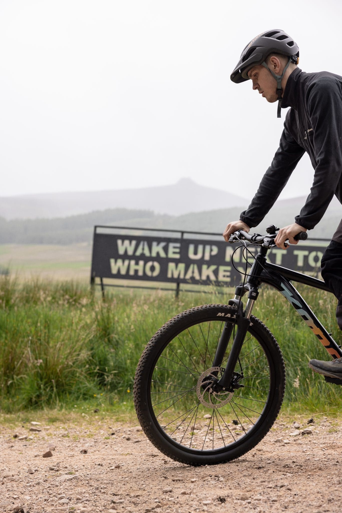 Mountain biker in helmet riding a gravel trail at Glen Dye, misty Scottish hills and a bold motivational sign visible behind