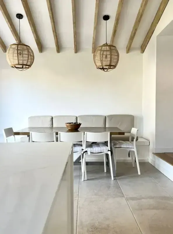 Airy holiday let dining area with built-in bench seating, wooden beam ceiling, and rattan pendant lights above a stone-toppe…