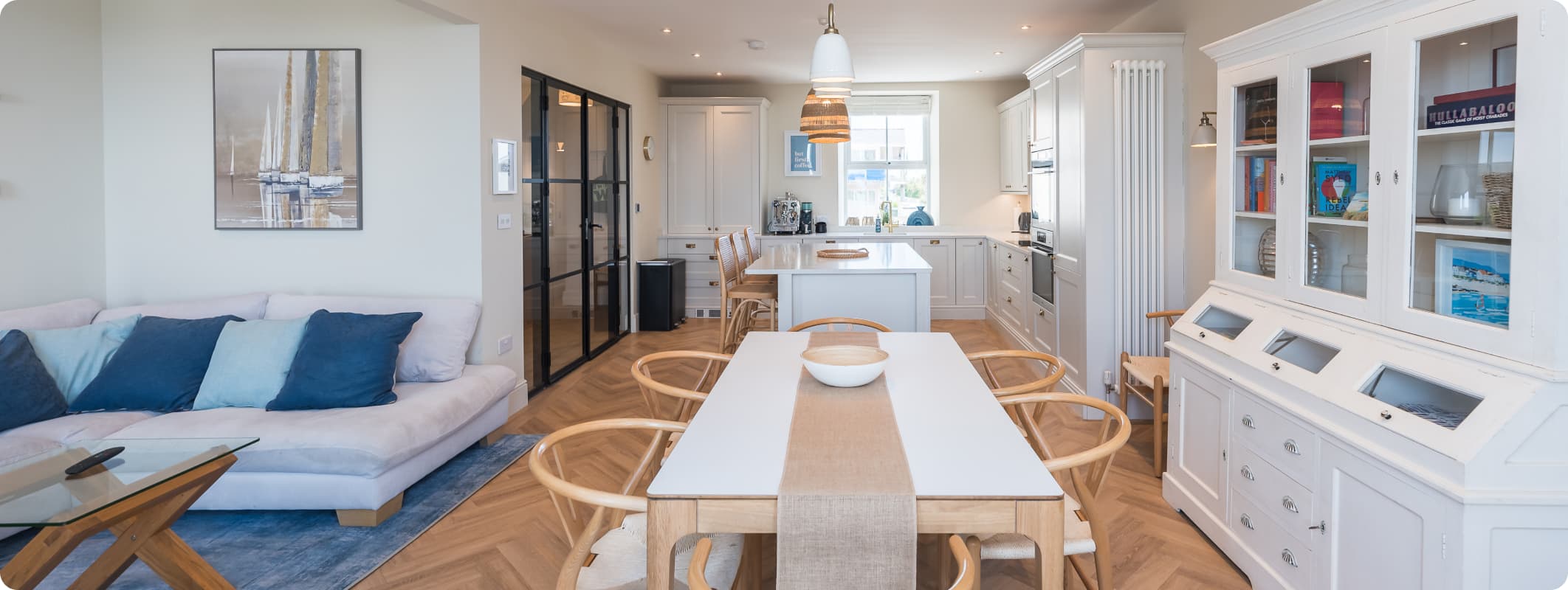 Open-plan kitchen, dining and living area in a luxury holiday cottage with herringbone oak floors, white shaker cabinetry, a…