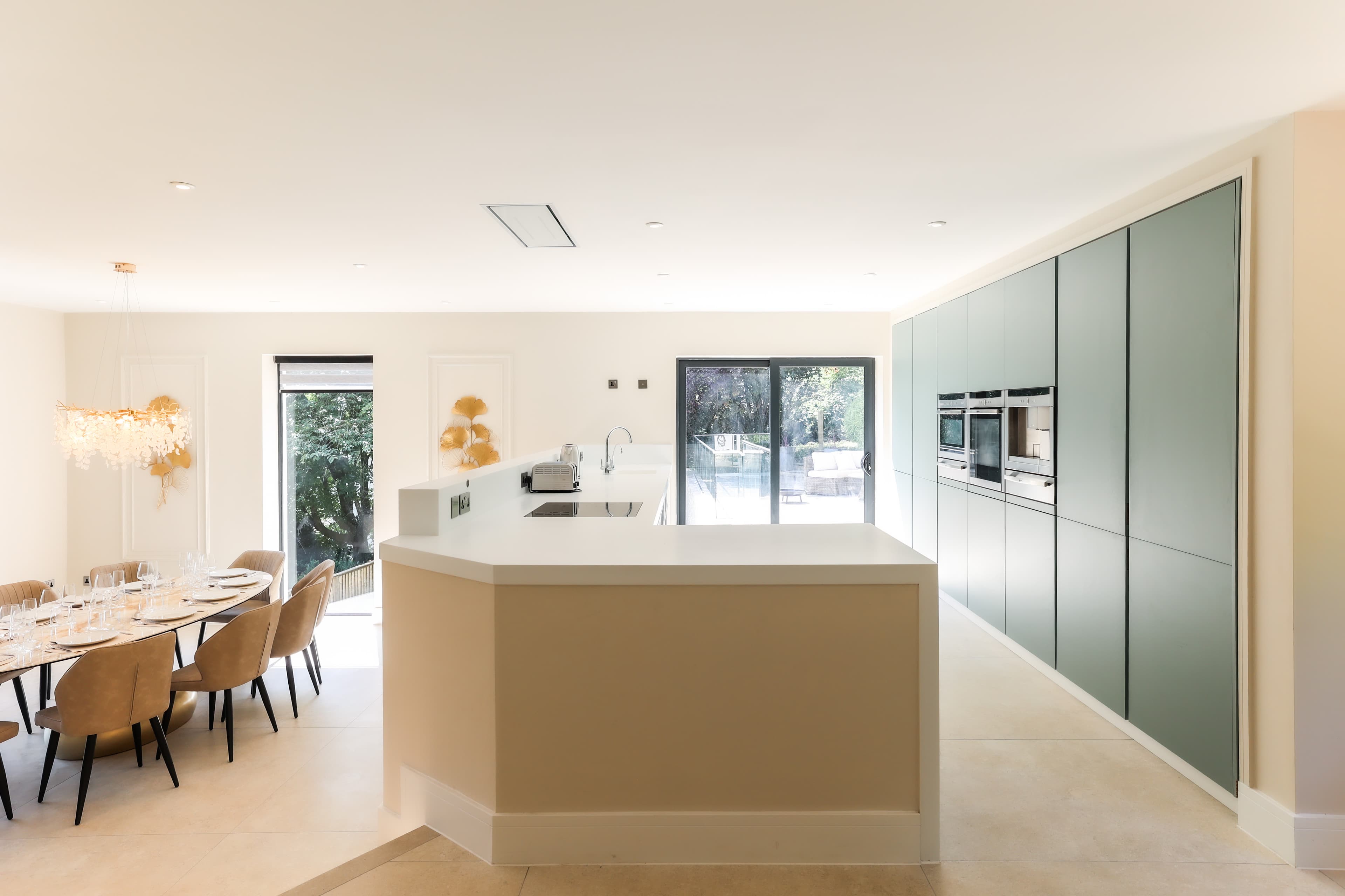 Open-plan luxury kitchen with large central island, sage green cabinetry, integrated ovens, and dining area set for guests w…