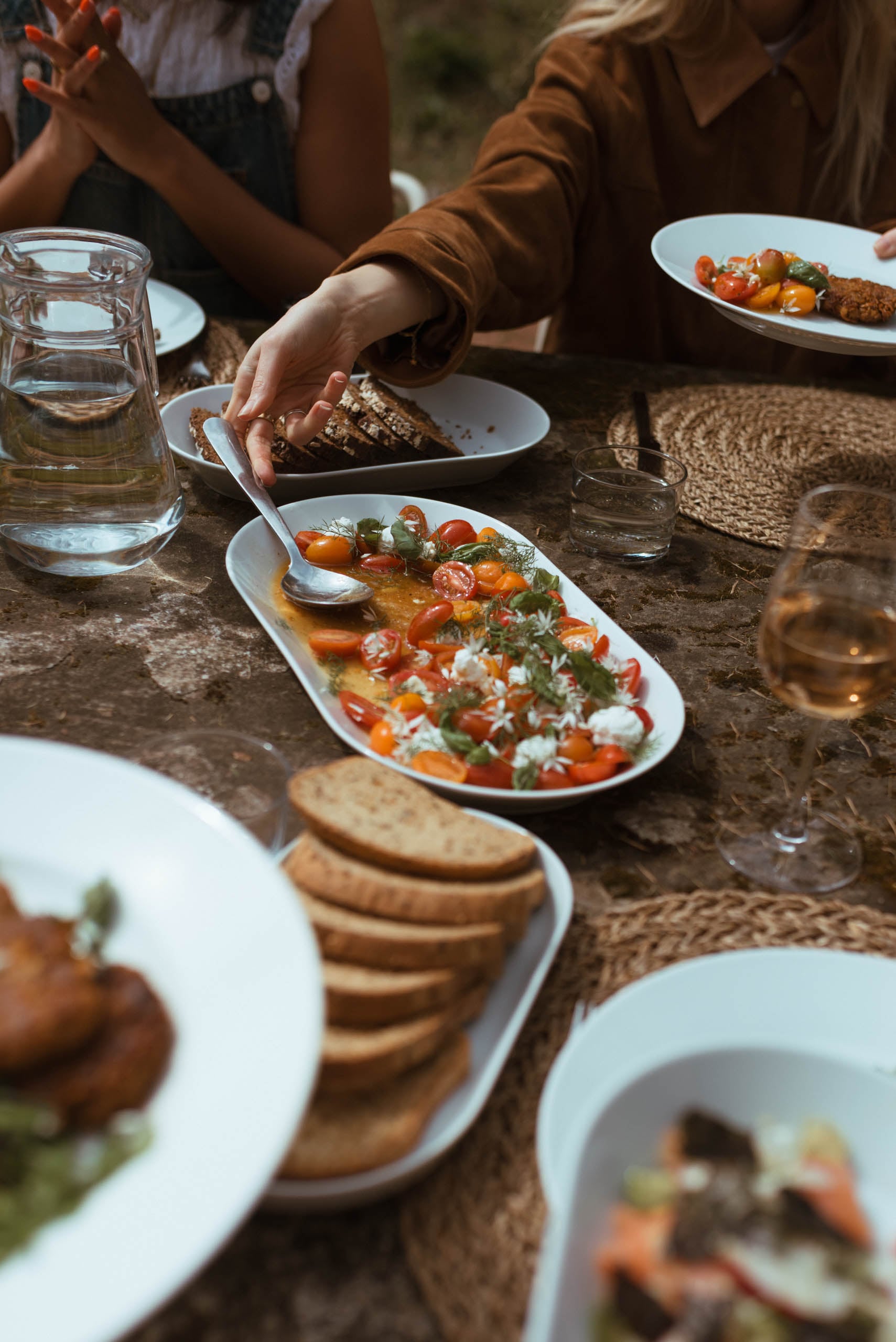 Guests sharing a rustic outdoor feast with a heirloom tomato and herb salad, artisan bread, and rosé wine on a stone table