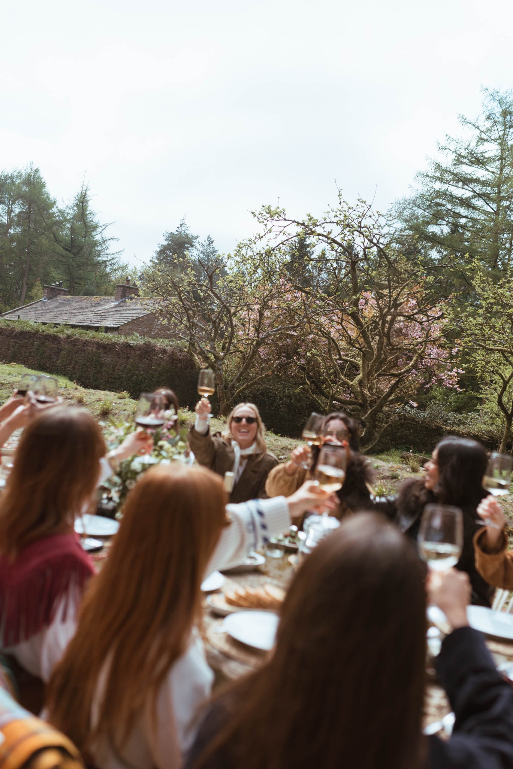 Group of women raising wine glasses in a toast at an outdoor long-table lunch in a walled garden, surrounded by spring bloss…