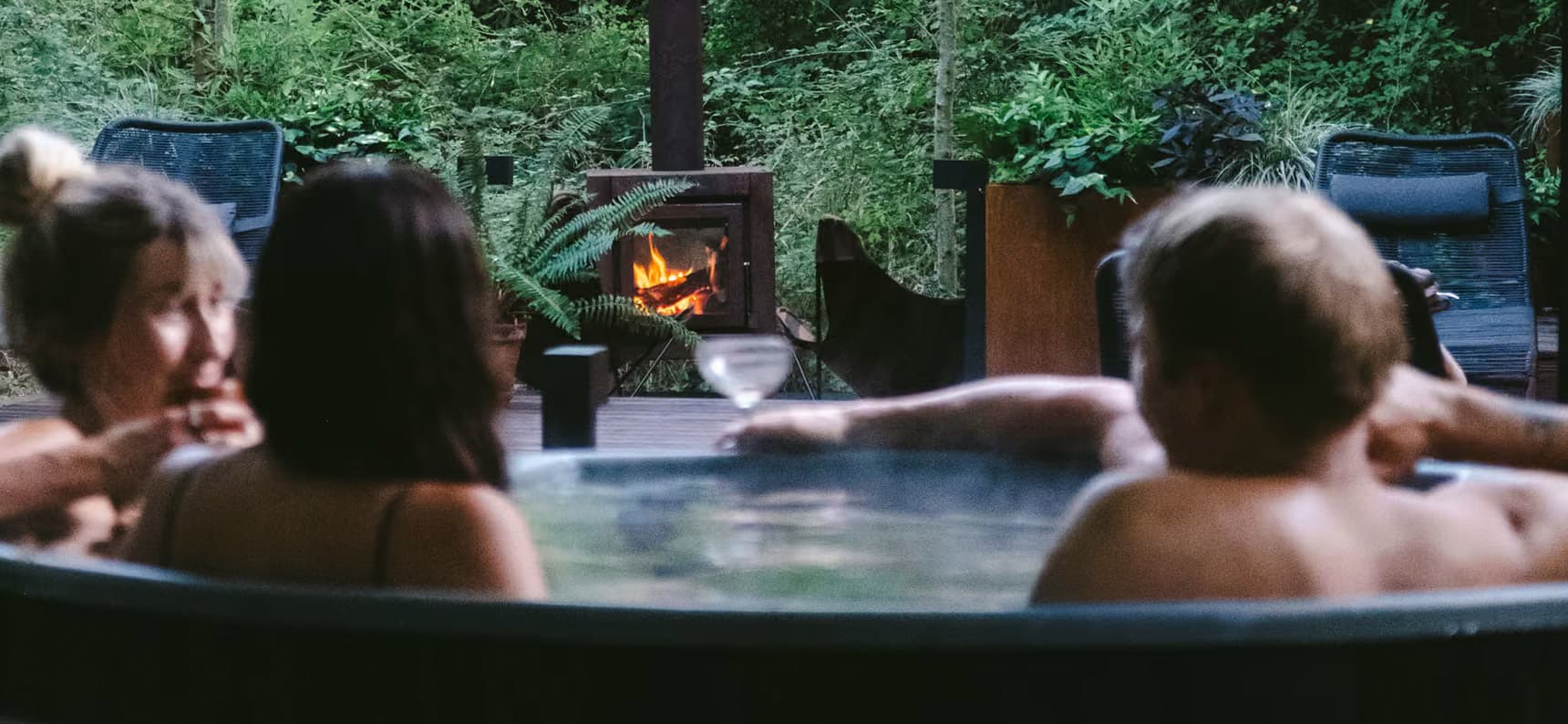 Guests relaxing in an outdoor hot tub with wine, facing a glowing wood burner stove surrounded by lush woodland