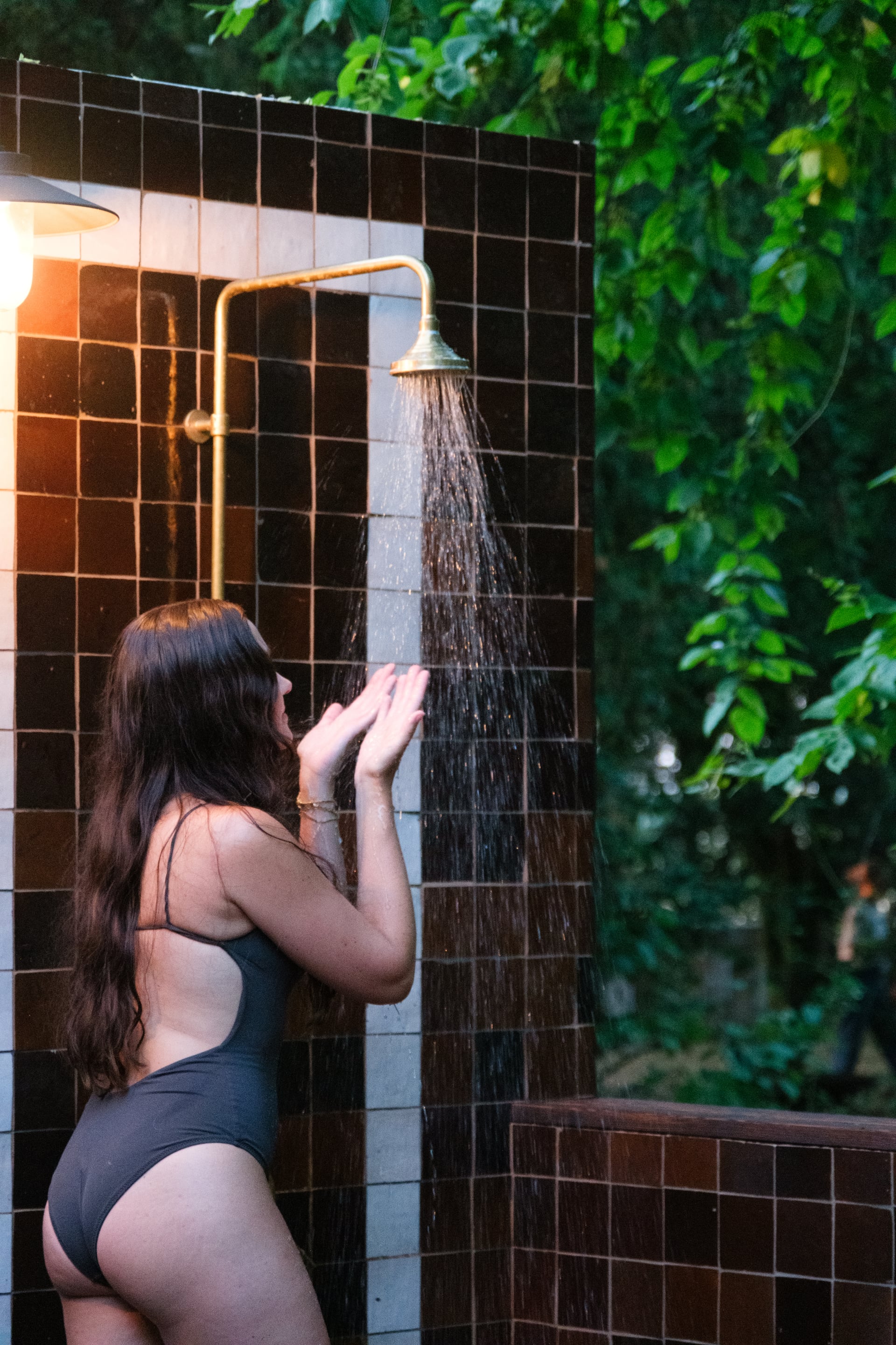Guest in swimsuit rinsing off under a brass outdoor shower head set against dark tiled wall surrounded by lush greenery