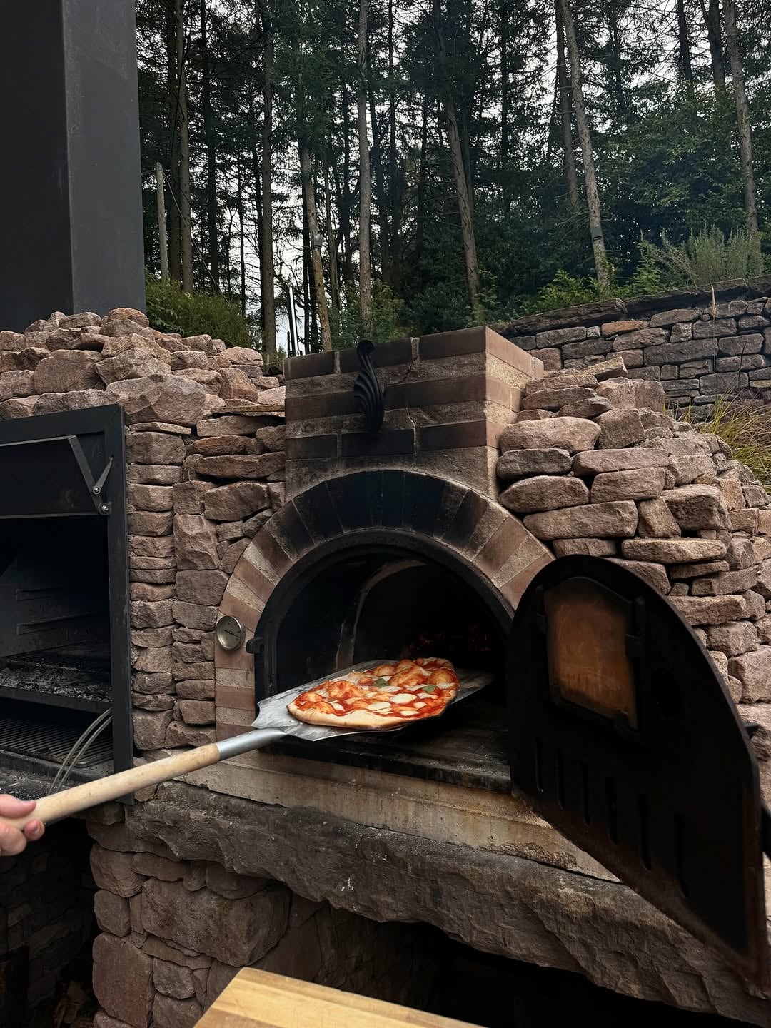 Freshly made margherita pizza being pulled from a stone-built outdoor wood-fired oven surrounded by woodland trees