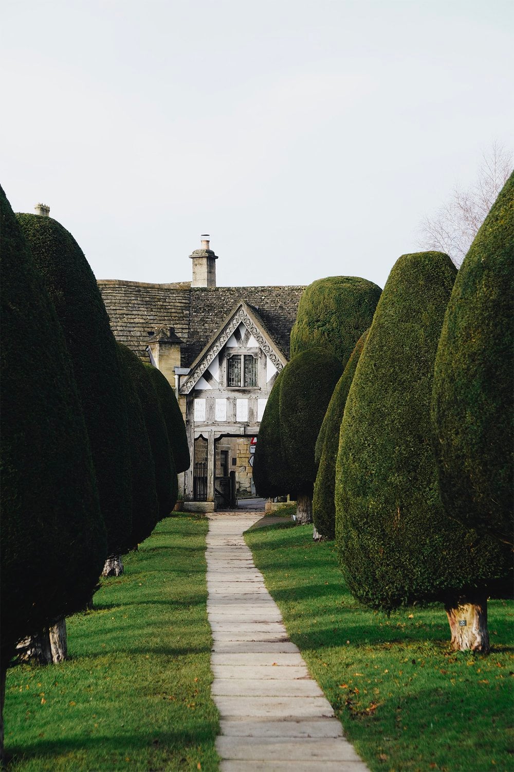 Stone pathway lined with ancient clipped yew trees leading to a historic Cotswold cottage with timber-framed gable in Painsw…