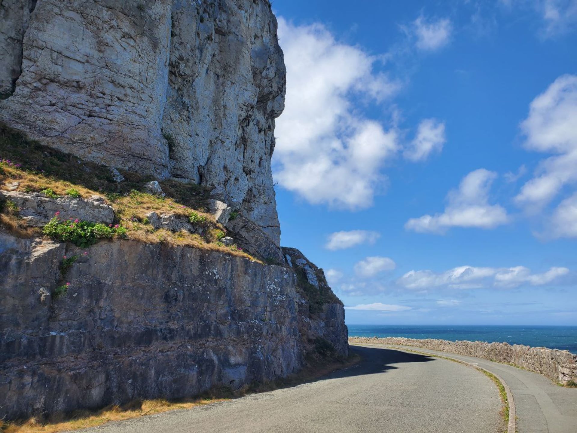 Narrow coastal road curving beneath dramatic limestone cliffs with the blue sea visible beyond a dry-stone wall, Great Orme,…