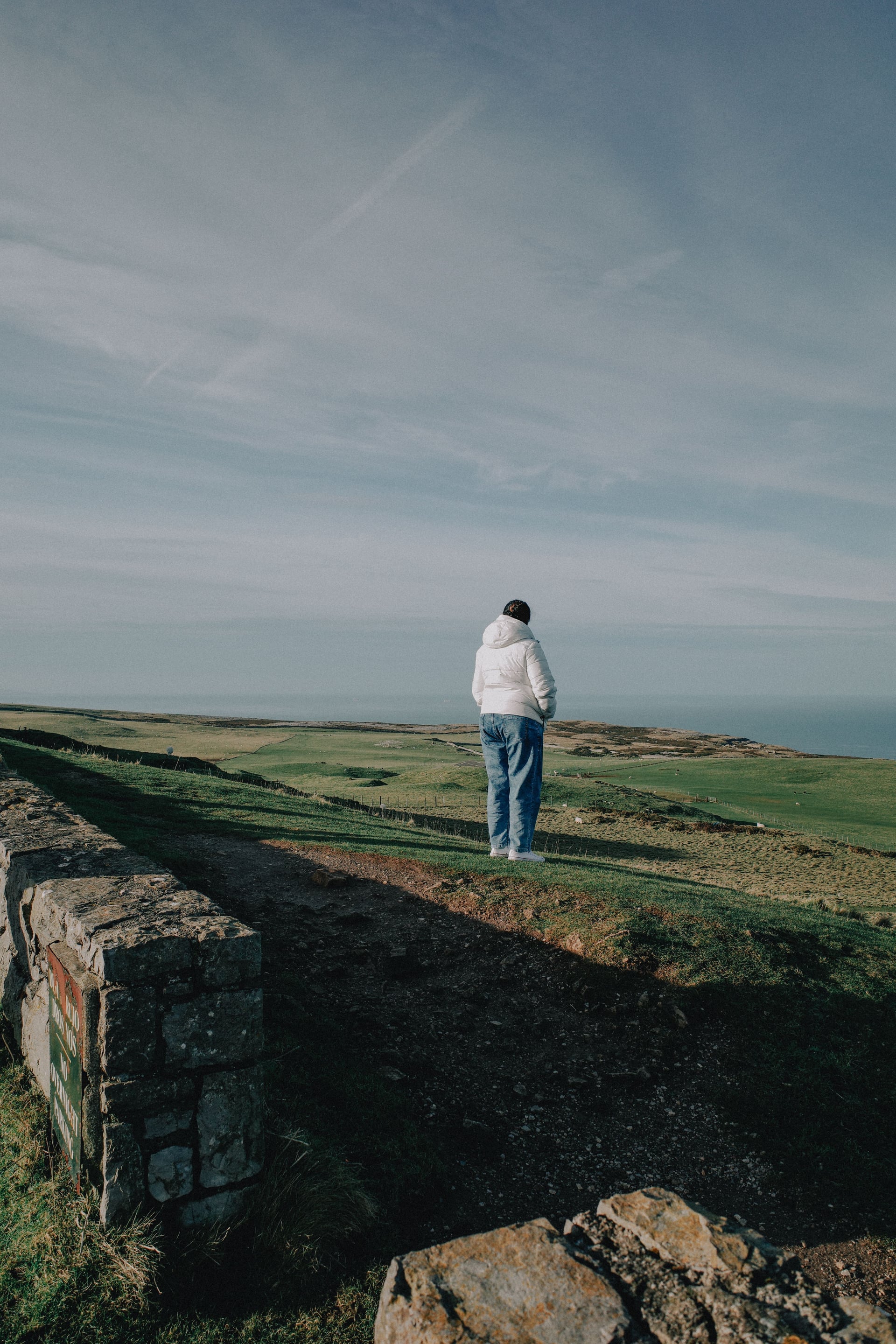 Guest in a white puffer jacket gazing across open coastal headland with rolling green fields and sea on the horizon