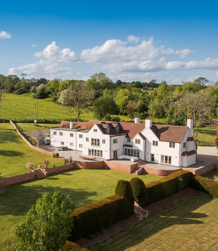 Aerial view of a large white country manor holiday let with terracotta roof, manicured lawns, and rolling Warwickshire count…