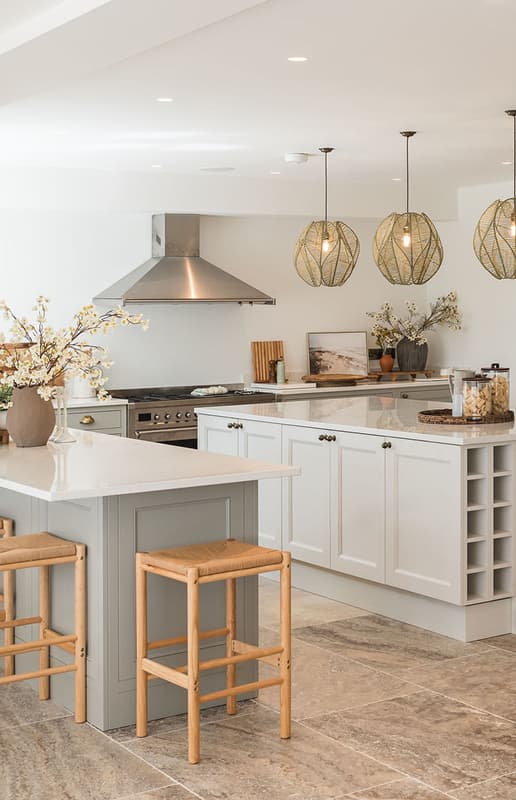 Bright open-plan kitchen with sage green island, rattan bar stools, woven pendant lights, and a range cooker beneath a steel…