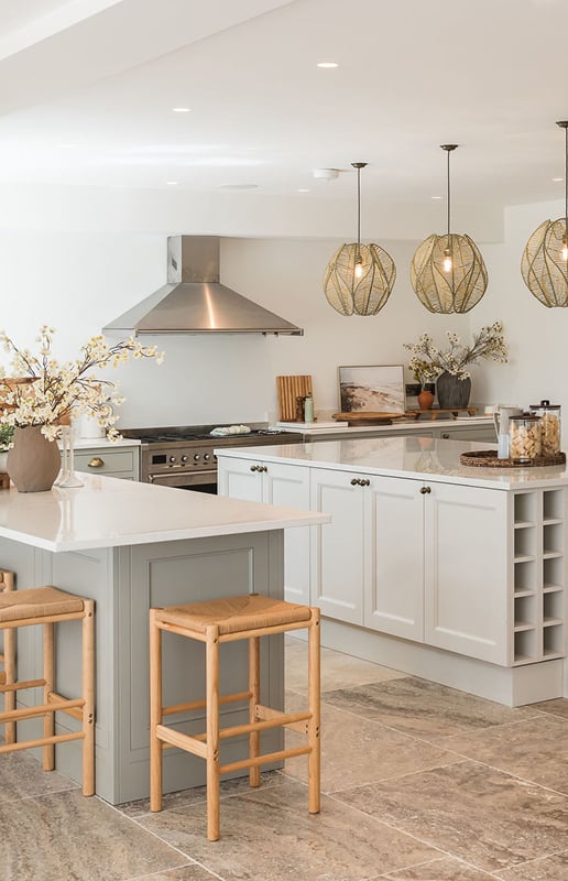 Bright open-plan kitchen with sage green island, rattan bar stools, woven pendant lights, and a range cooker beneath a steel…