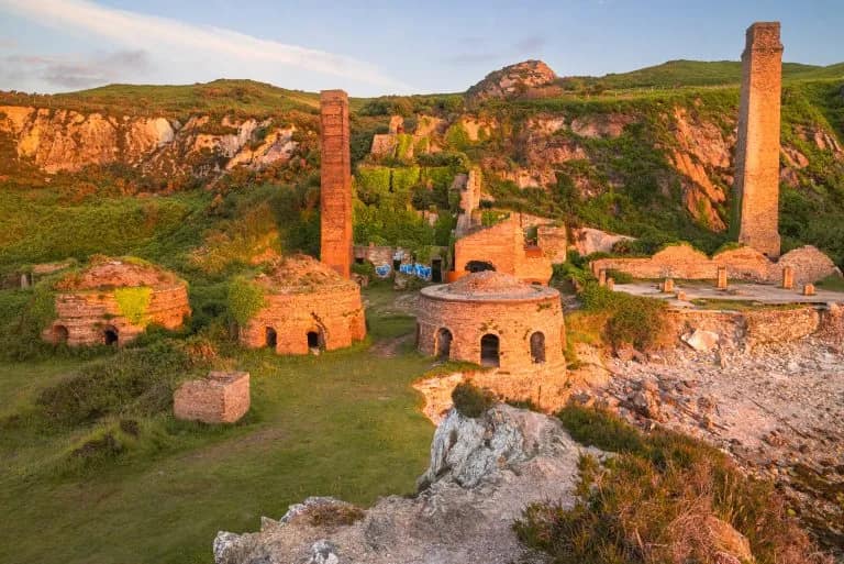 Ruins of Porth Wen brickworks on Anglesey at golden hour, with circular kilns and tall brick chimneys set against green coas…