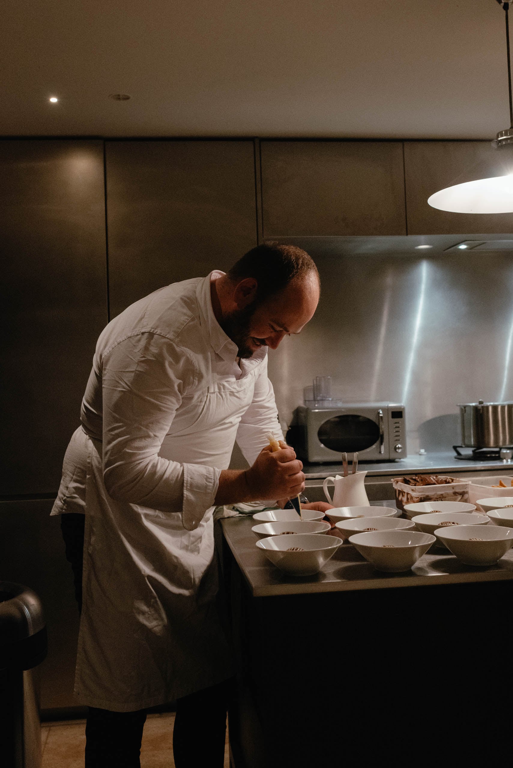 Private chef carefully plating multiple bowls in a sleek dark kitchen, showcasing the in-house chef experience available at…