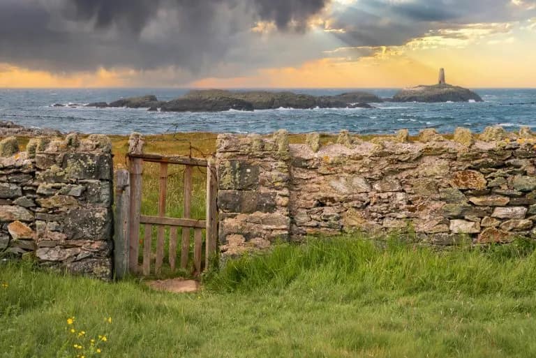 Weathered dry stone wall and wooden gate opening onto a dramatic Anglesey coastline at sunset, with a small island and monum…