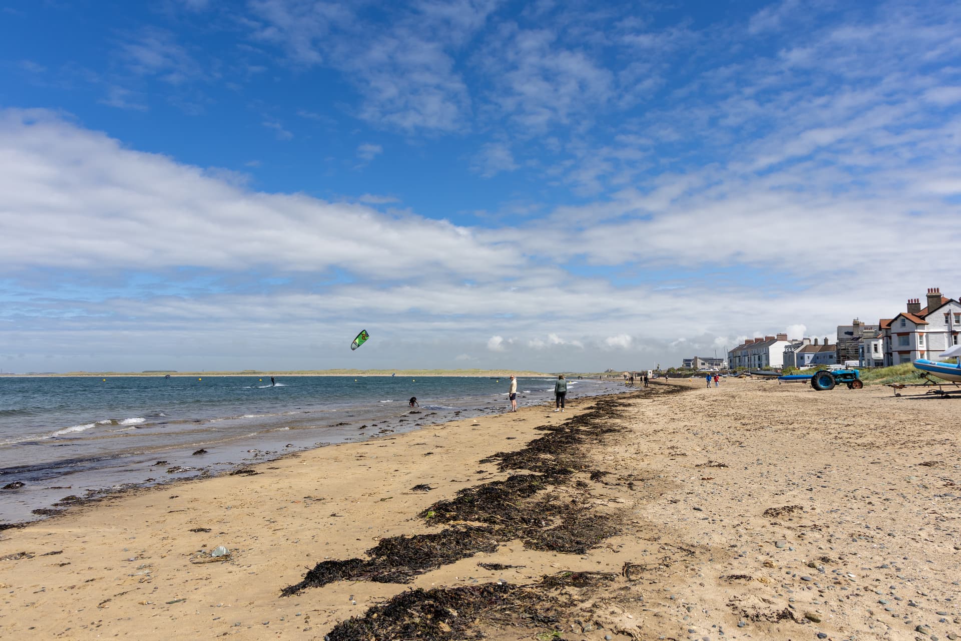 Sandy beach with a kite surfer on the water, dog walkers strolling the shoreline, and coastal cottages lining the seafront u…