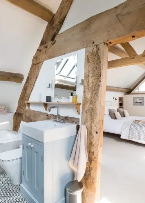 Open-plan en suite bathroom with exposed oak beams, grey vanity unit, and a double bedroom beyond in a converted cottage