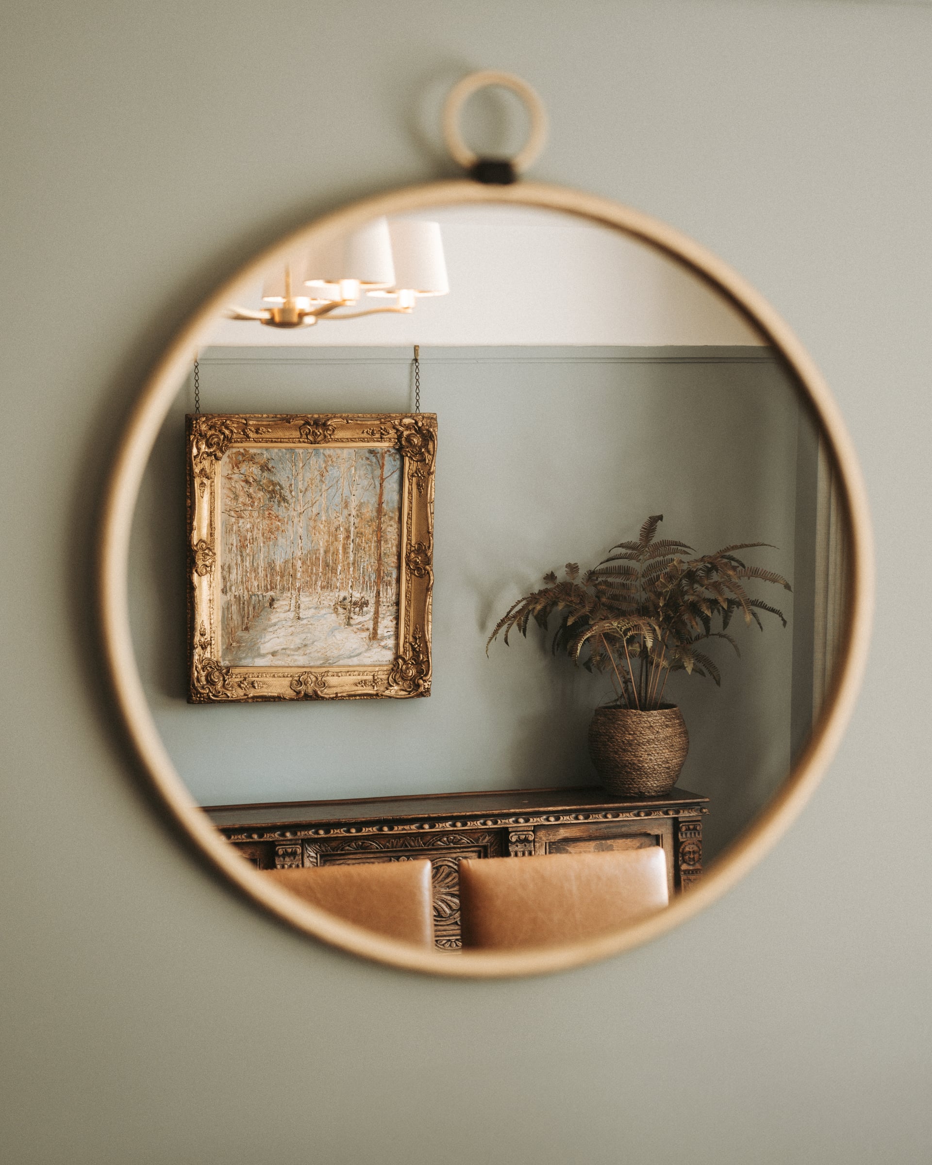 Round gold-framed mirror reflecting an ornate gilt painting, carved console table, woven plant pot, and brass chandelier in…