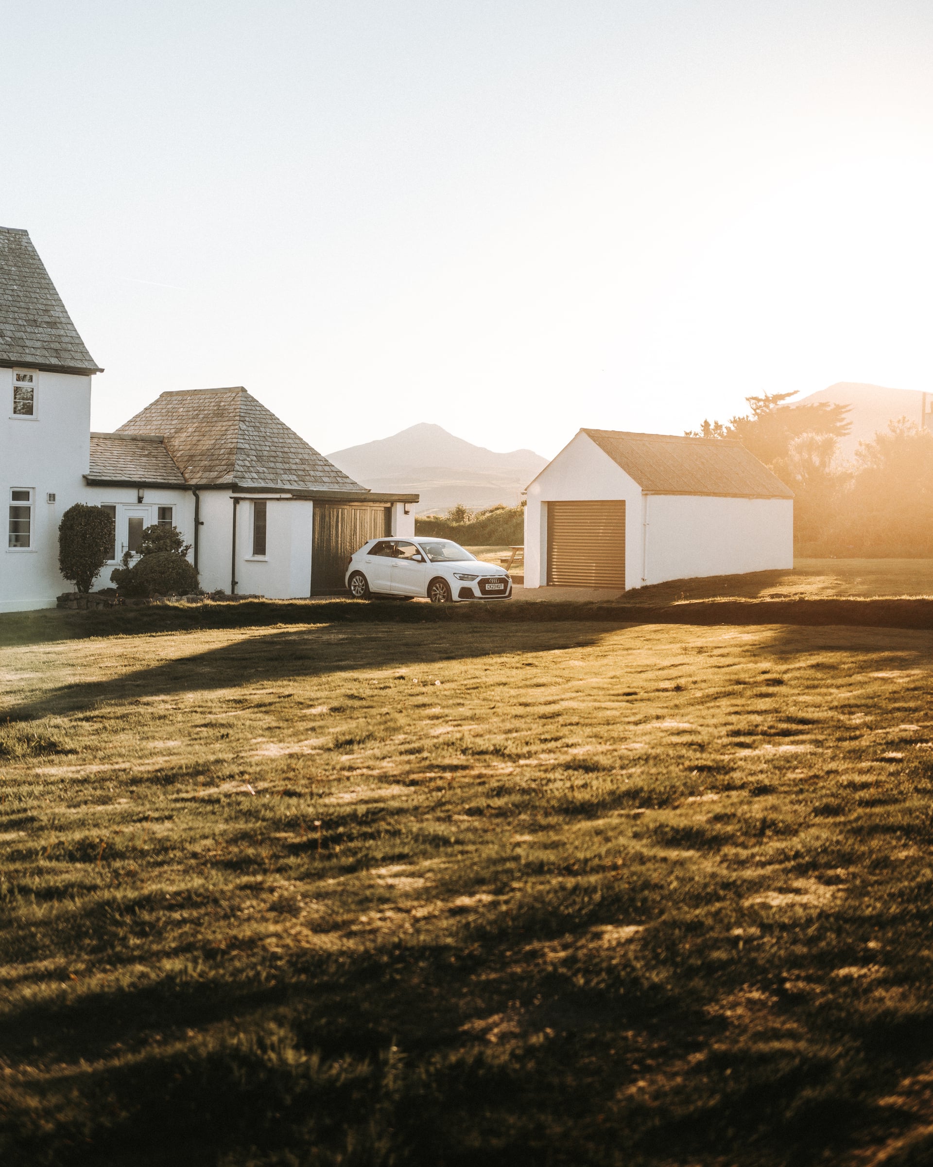 White Scottish holiday cottage with private parking and outbuildings bathed in golden sunset light, mountains visible in the…
