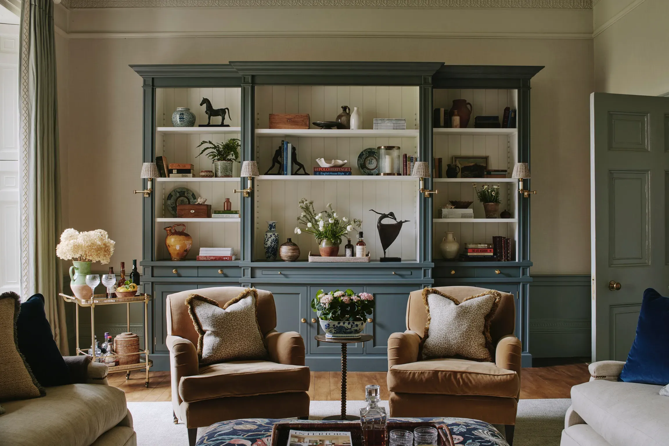 Elegant sitting room with a large slate-blue fitted bookcase styled with ceramics, books, and fresh flowers, flanked by two…