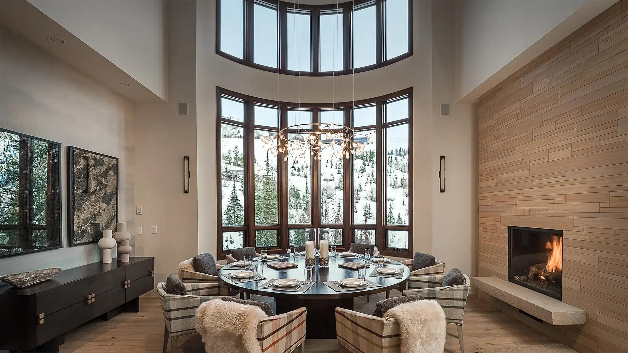 Luxury ski chalet dining room with curved floor-to-ceiling windows overlooking snowy mountain slopes, lit fireplace, and rou…