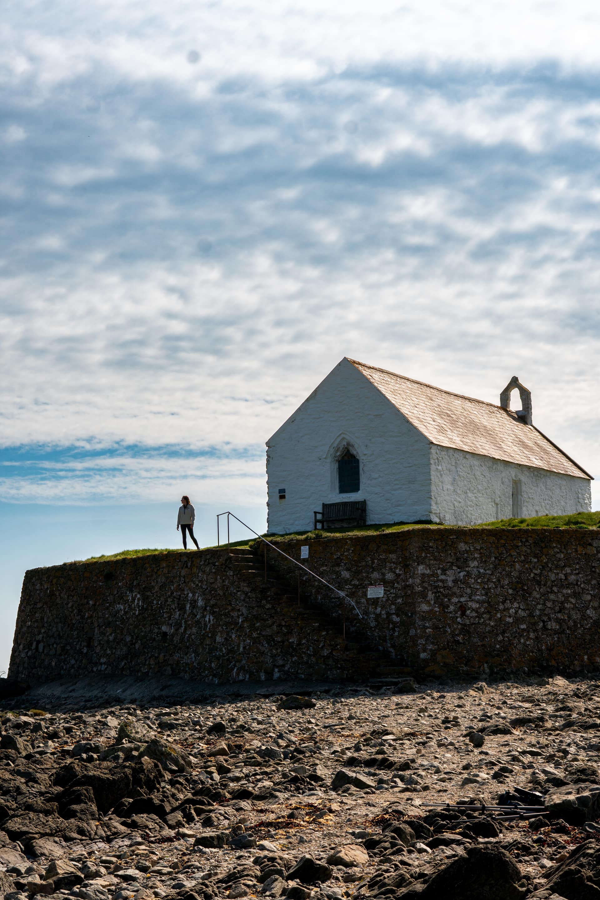 Walker beside the whitewashed medieval St Cwyfan's Church perched on a rocky tidal island off the Anglesey coastline, Wales