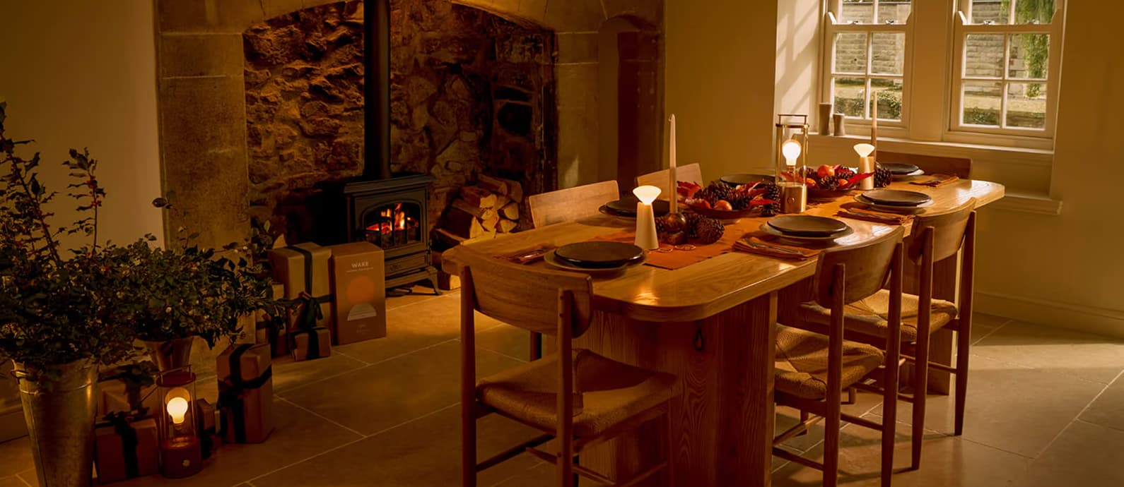 Candlelit dining room in a stone cottage with a roaring log burner, rustic oak table set for six guests with seasonal autumn…