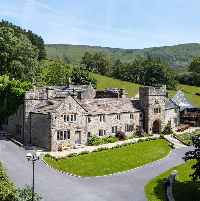 Aerial view of a grand stone manor house nestled in a green Peak District valley, with manicured lawns, a gatehouse tower, a…