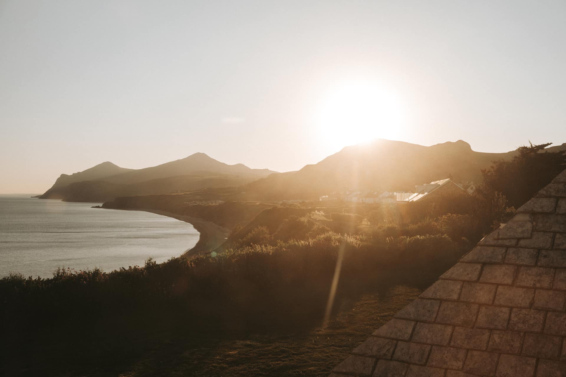 Golden sunset over a sweeping Welsh bay with mountain headlands and a curved sandy beach viewed from a hillside cottage terr…