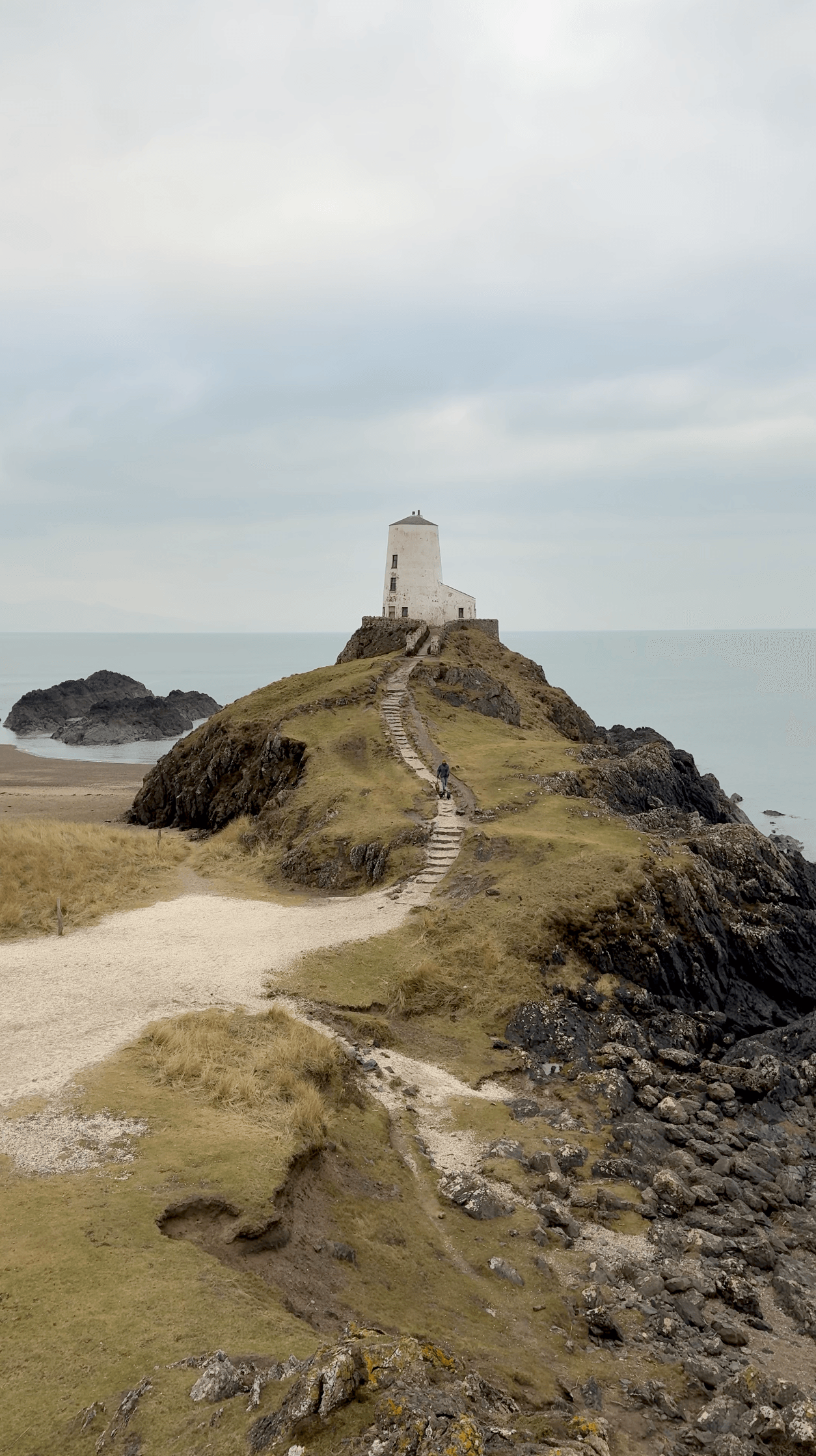 Stone path leading up to Tŵr Mawr lighthouse on Llanddwyn Island, Anglesey, with a walker and calm Irish Sea beyond