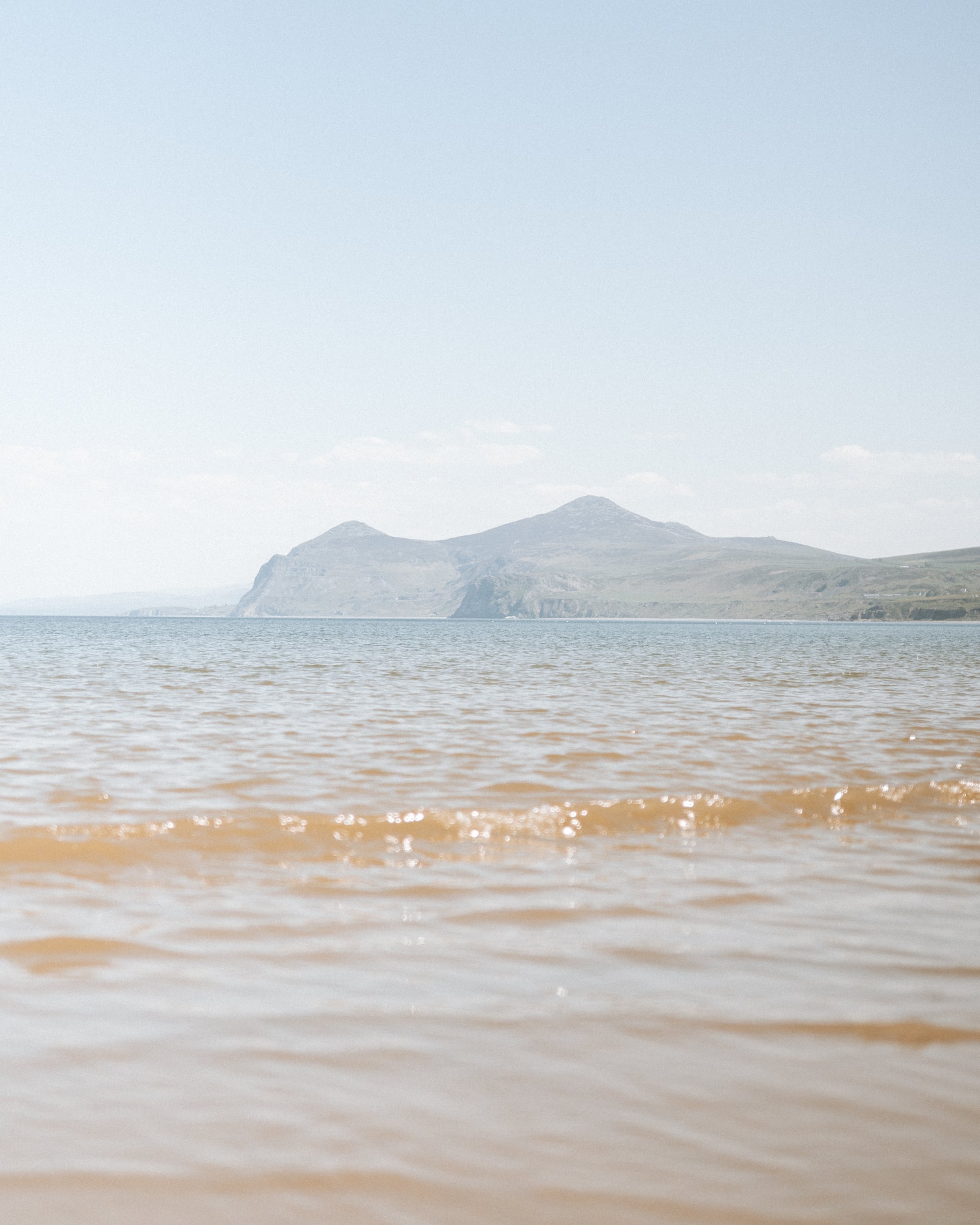 Shallow golden shoreline with gentle waves and a dramatic mountain headland rising across a calm bay on the Welsh coast
