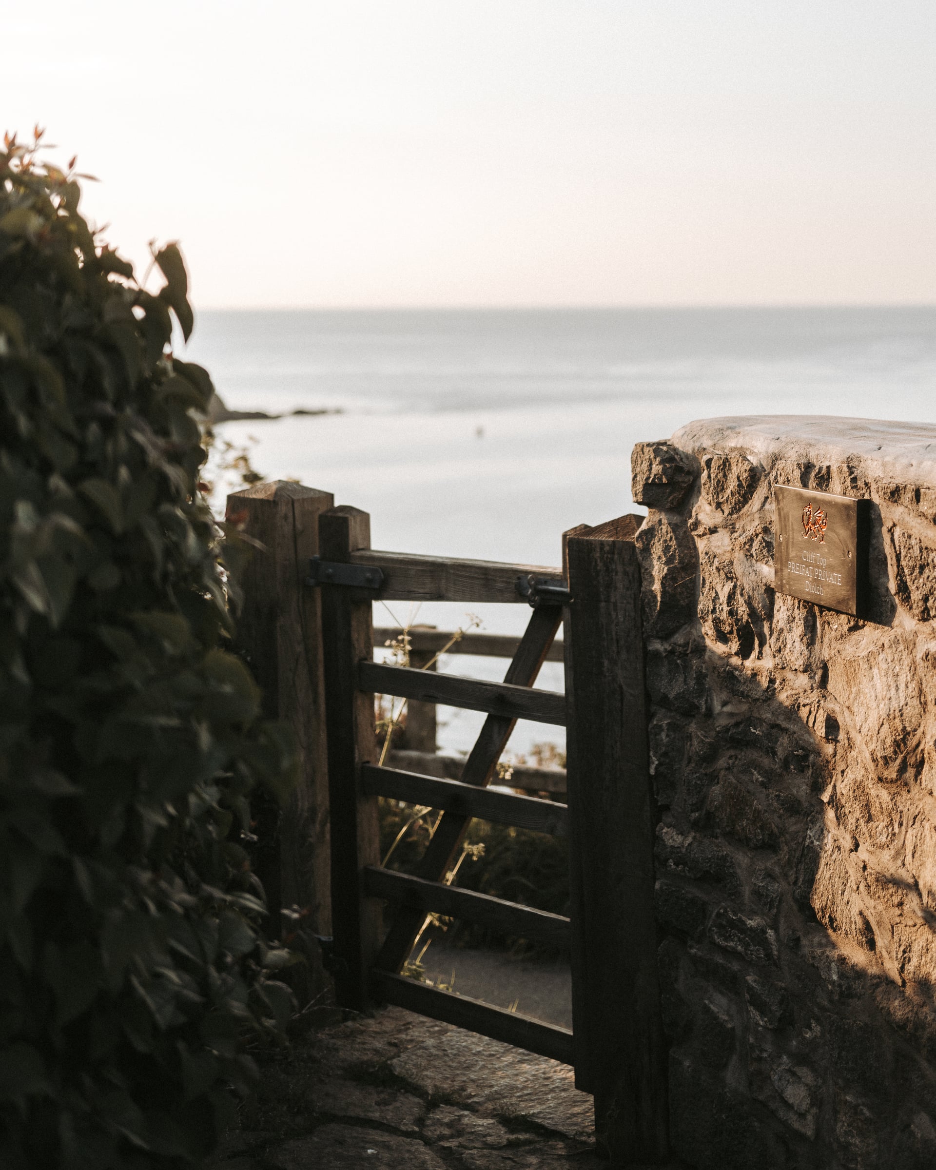 Rustic wooden gate set in a stone wall opening onto a coastal path with calm sea views beyond, Wales