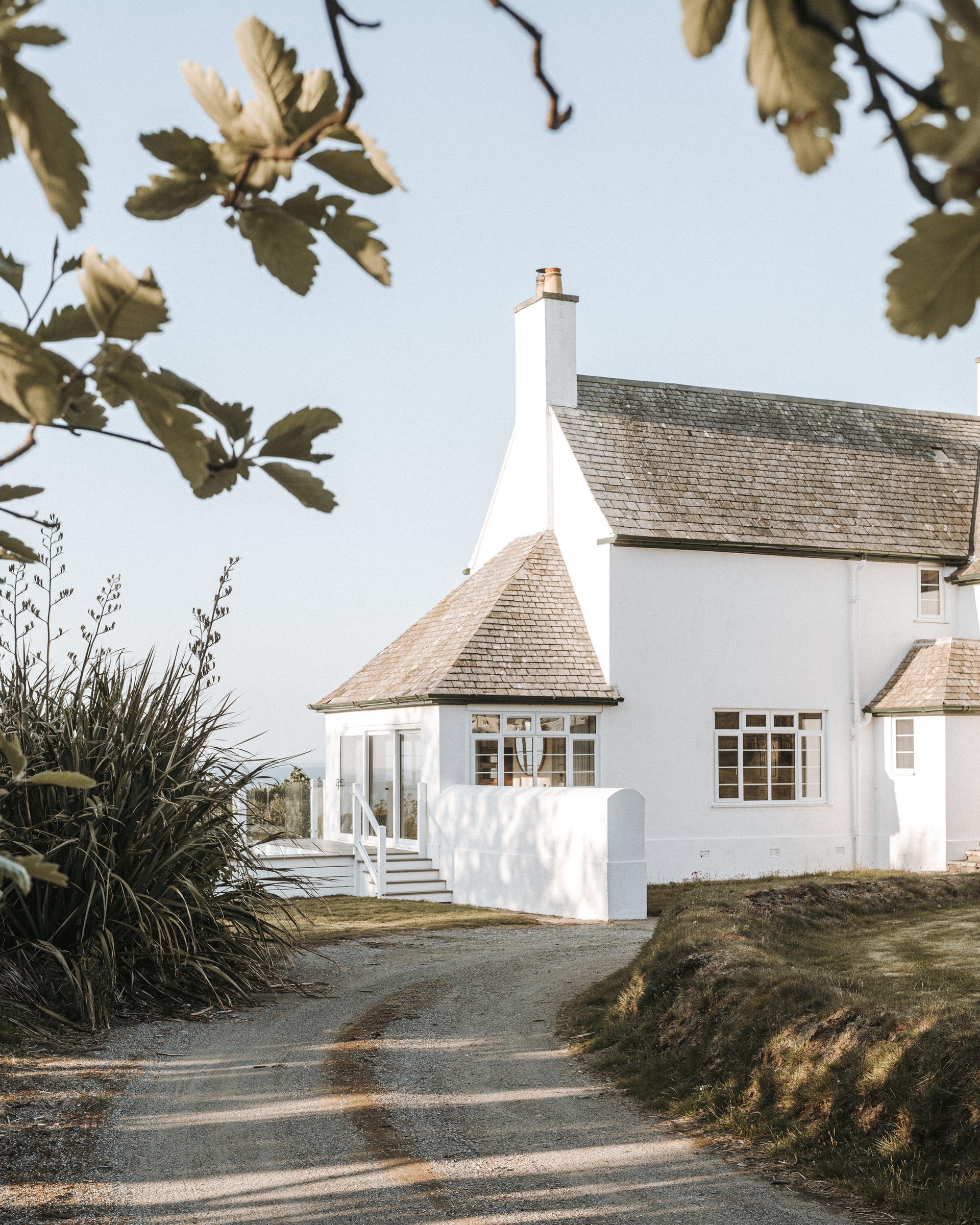Whitewashed coastal holiday cottage with slate roof, chimney, and glazed porch framed by windswept grasses and oak branches…