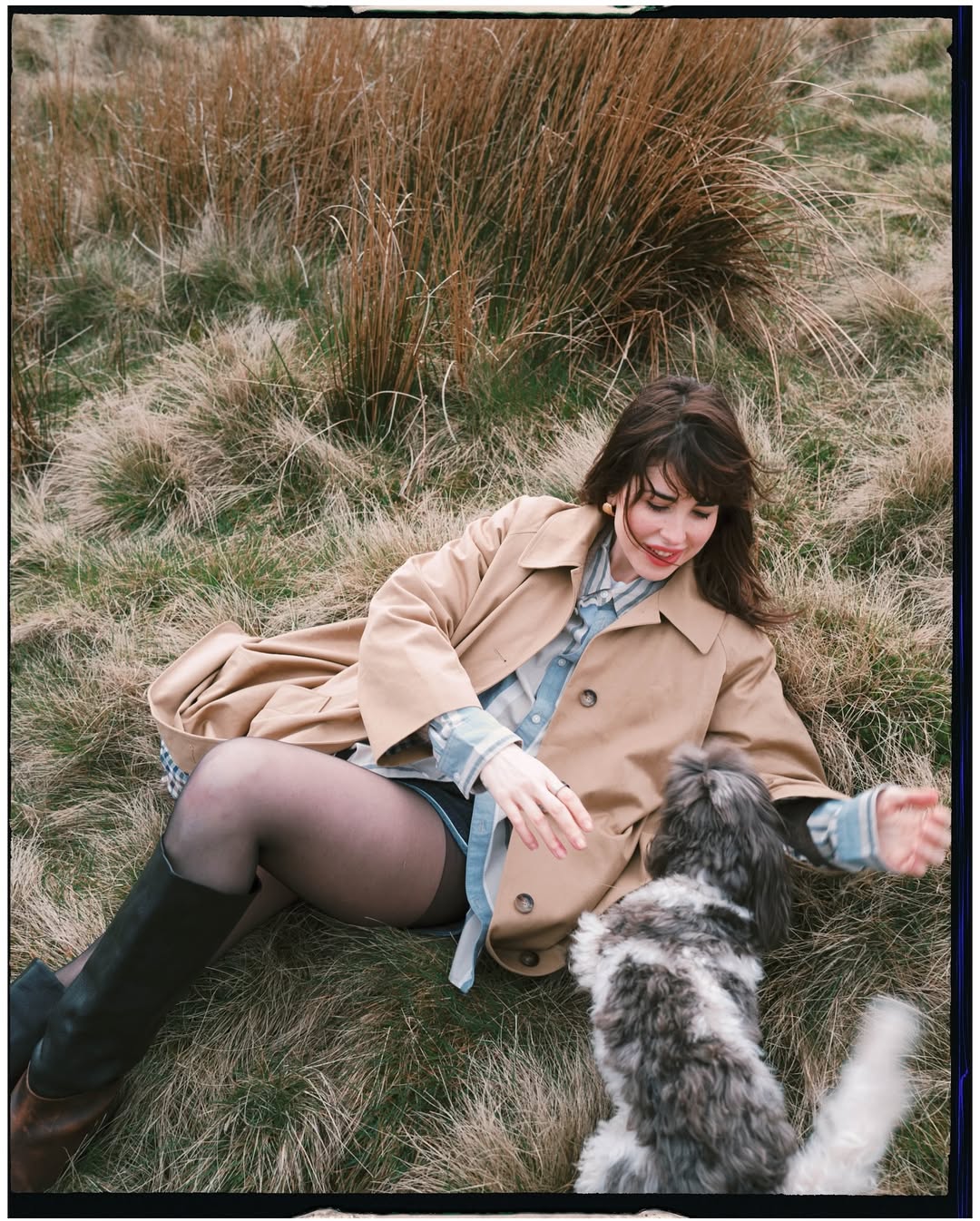 Woman in a trench coat and knee-high boots sitting on moorland grass, laughing as she plays with a fluffy dog on a countrysi…
