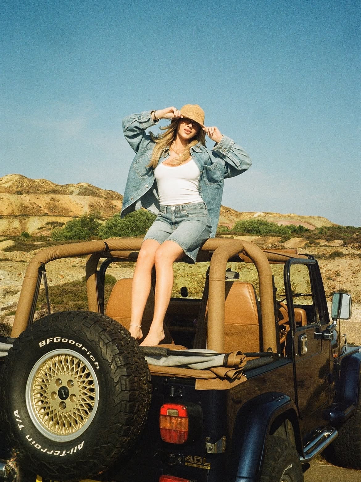 Woman in denim and straw hat perched on an open-top 4x4 Jeep against a sun-baked hillside landscape, evoking a carefree holi…