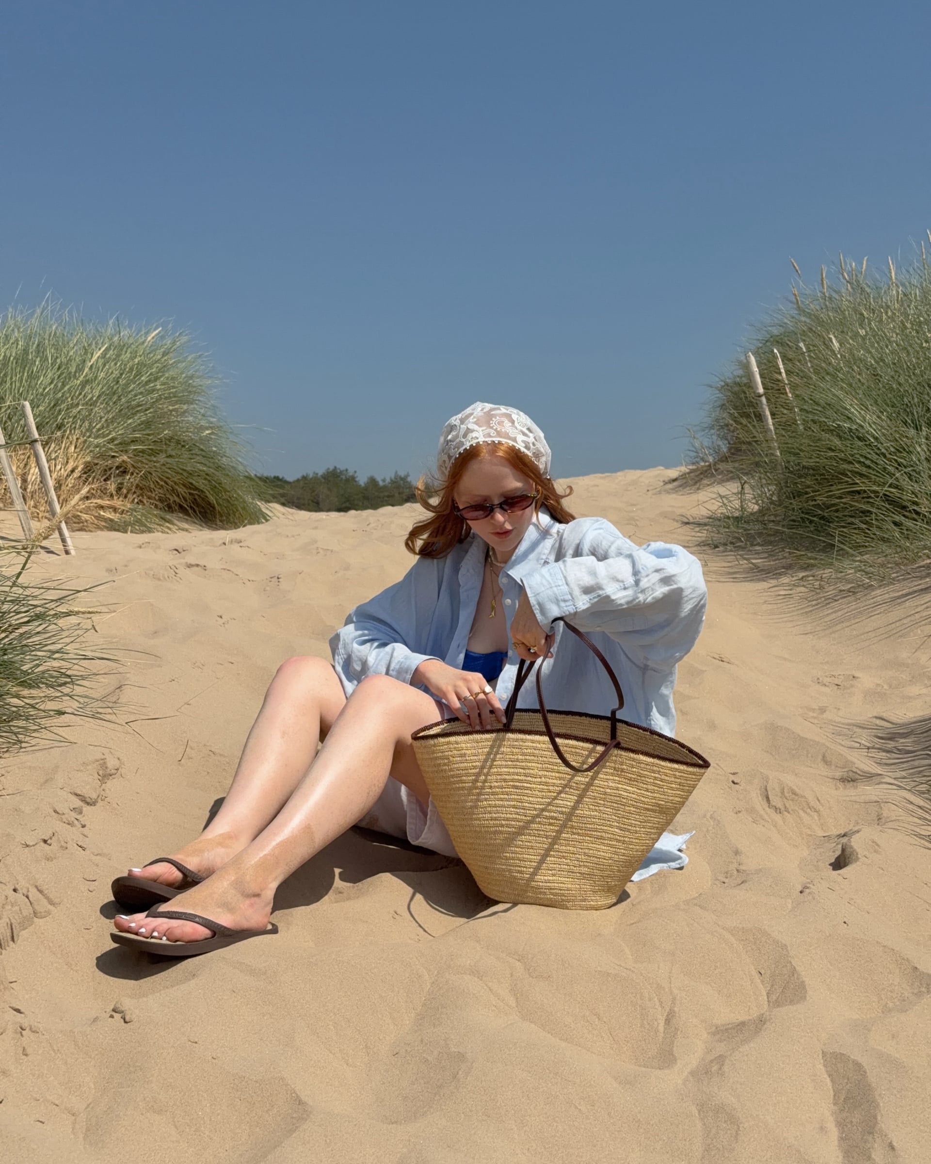 Woman relaxing on sandy dunes with a straw beach bag, linen shirt and lace headscarf on a sunny UK coastline