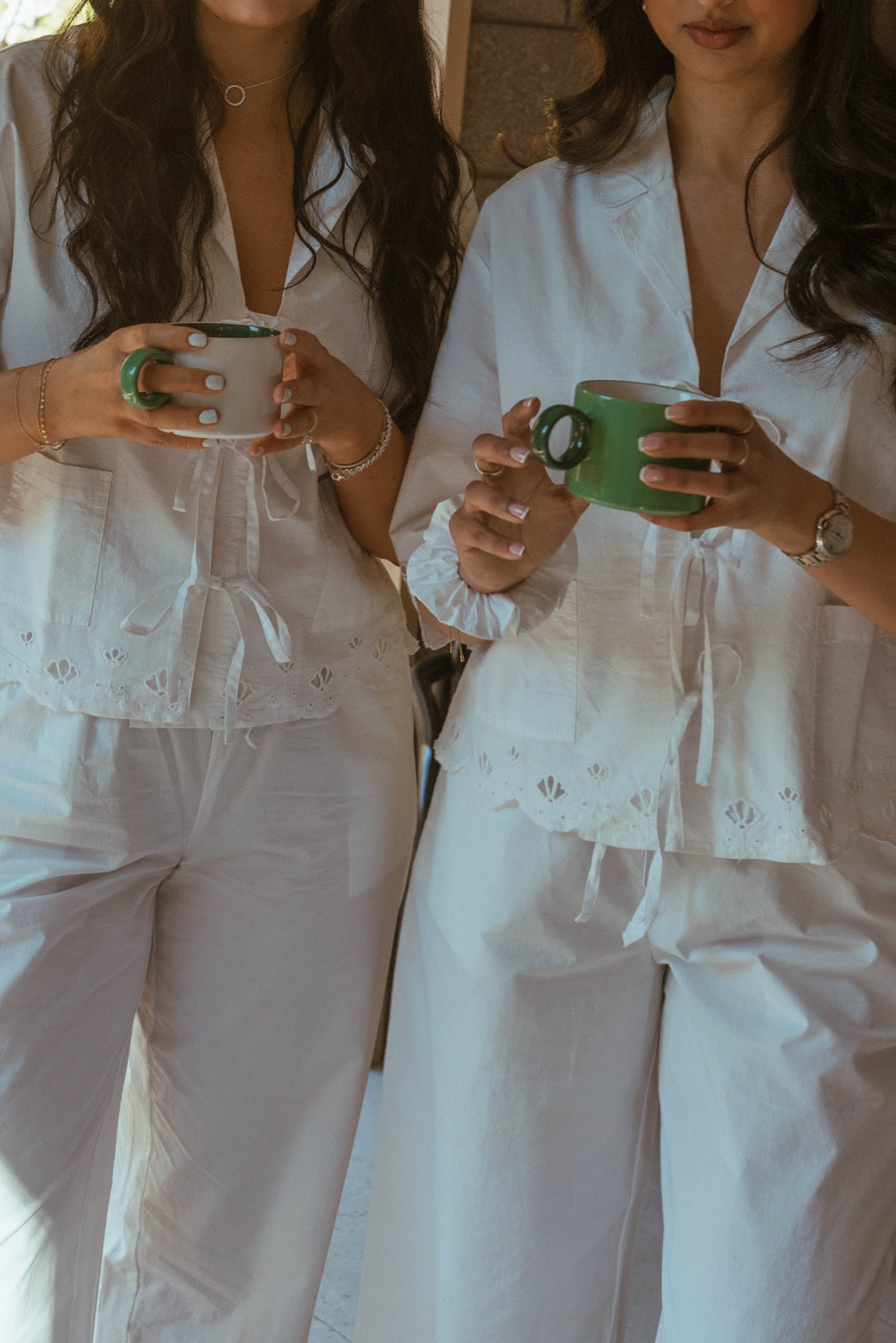 Two women in white broderie anglaise pyjamas holding green ceramic mugs, enjoying a relaxed morning coffee during a holiday…