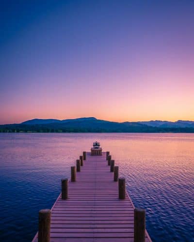 Wooden jetty stretching over a calm lake at sunset with purple skies and mountain silhouettes, Lake District