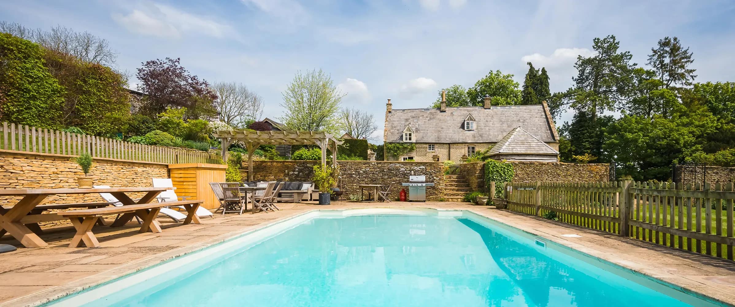 Outdoor heated swimming pool at a Cotswolds stone holiday cottage, with wooden dining furniture, pergola, and BBQ on a sunny…
