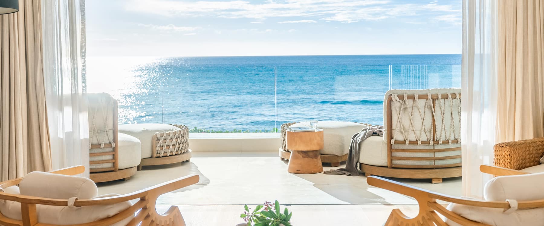 Bright open-plan living area with rope-woven daybeds and floor-to-ceiling glass doors opening onto a stunning direct sea view