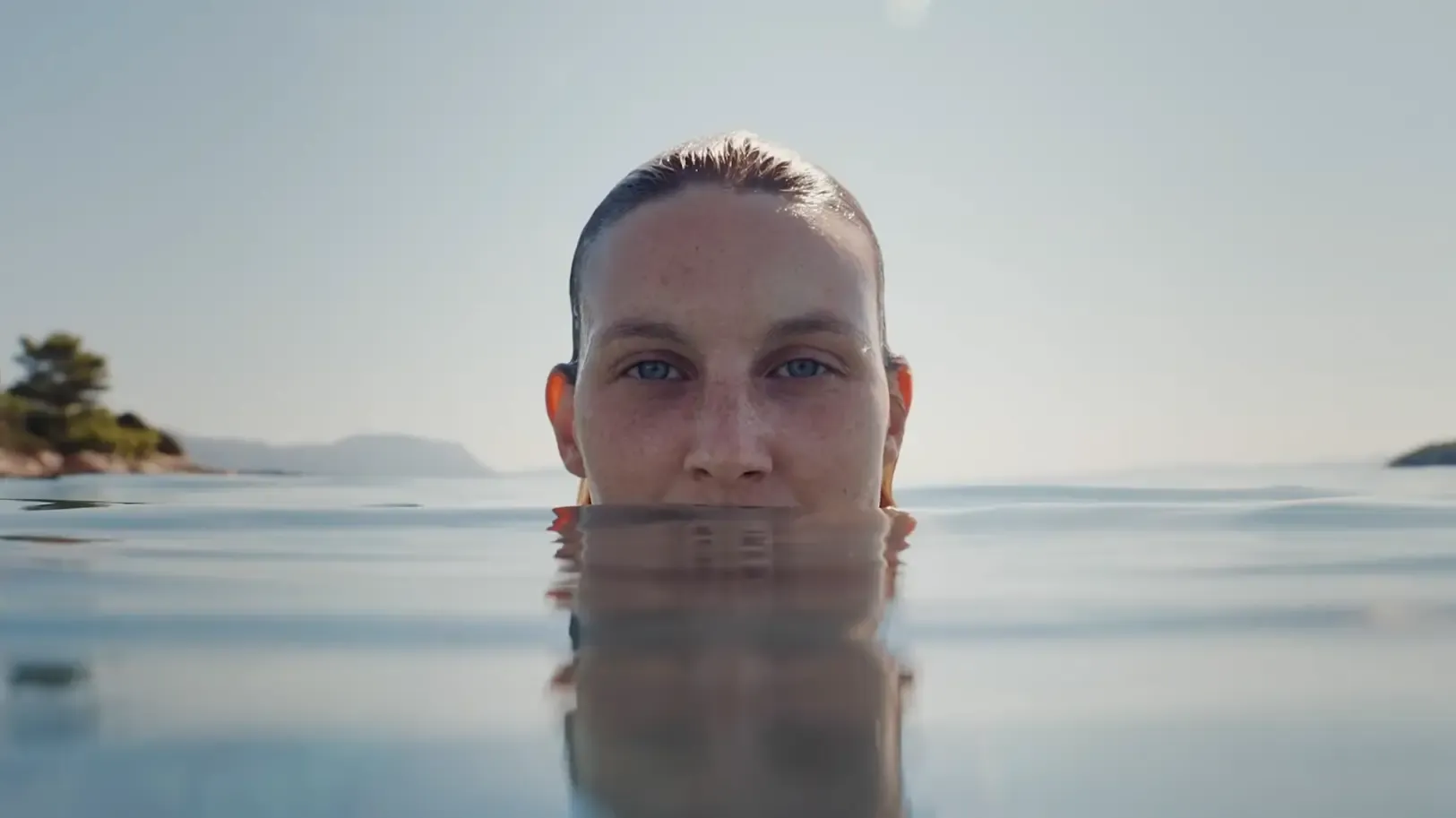 Woman floating at water level in a calm sea with wooded islands and hazy coastal hills in the background